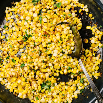 A silver serving spoon tucked into a skillet of sautéed corn, garnished with fresh basil.