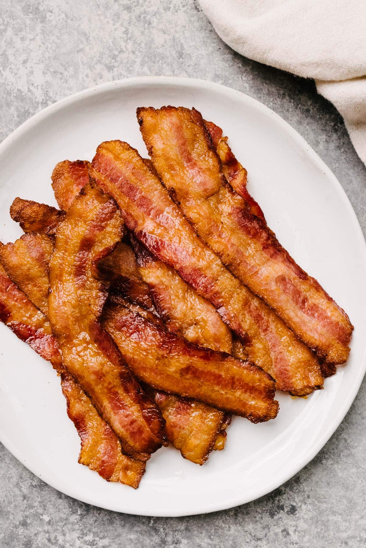 Oven cooked bacon strips on a white plate with tan linen napkin to the side.