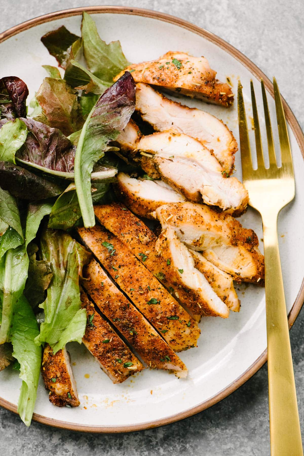 Thinly sliced pieces of roasted boneless chicken thighs on a plate with a tossed green salad and a gold fork.