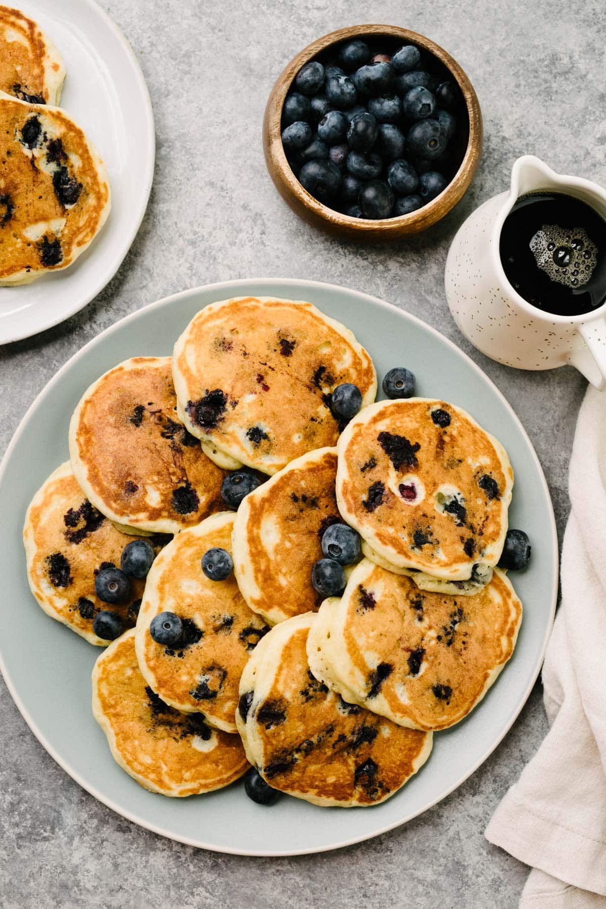 A platter of homemade blueberry pancakes with buttermilk on a concrete background, surrounded by a linen napkin, small pitcher of maple syrup, and bowl of fresh blueberries.
