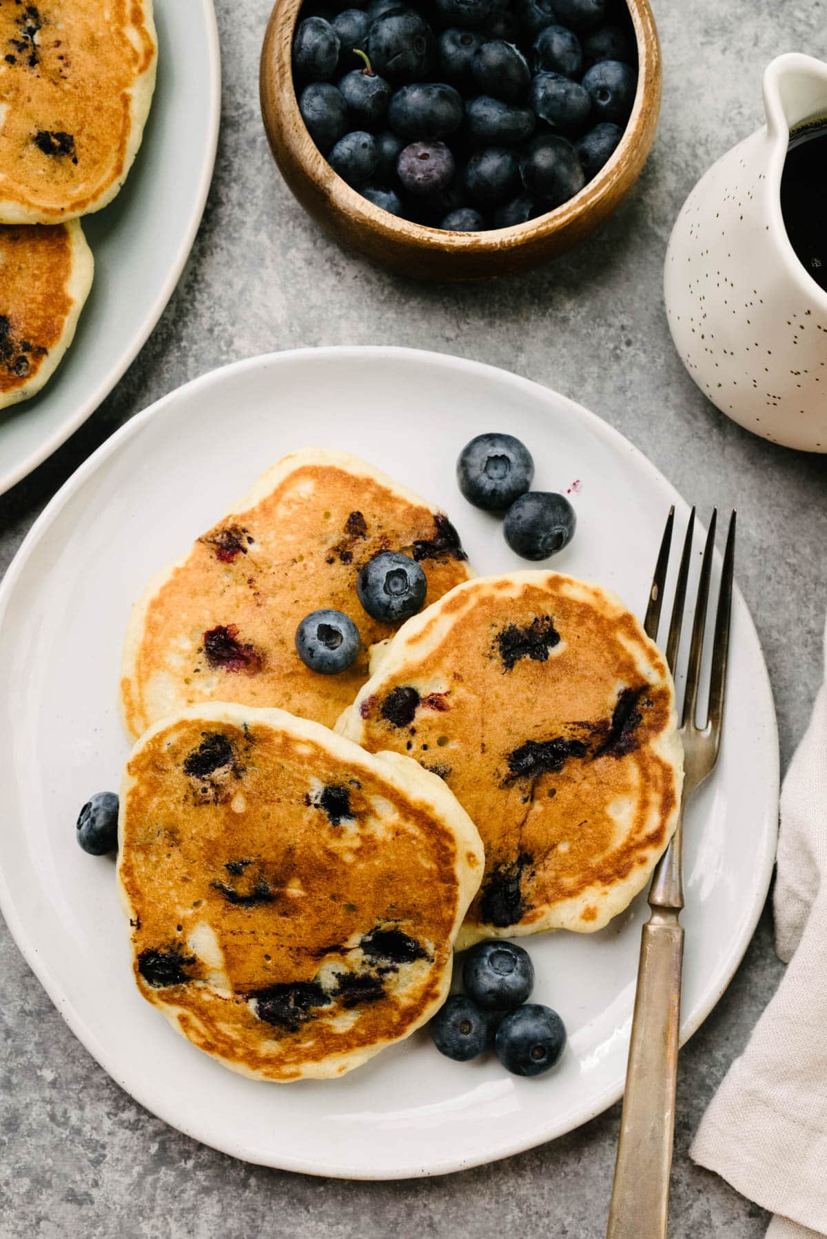Three blueberry pancakes with buttermilk on a white plate with a silver fork and additional fresh blueberries; plate is surrounded by a cream linen napkin, small pitcher of maple syrup, and a bowl of blueberries.