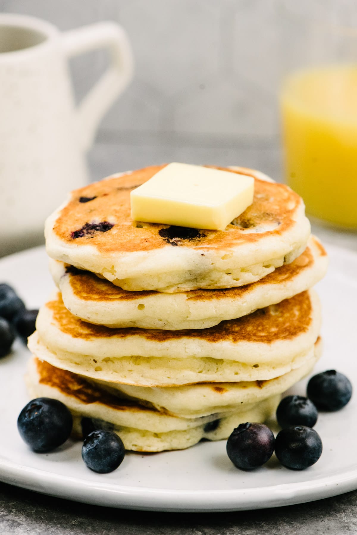 Side view, a stack of blueberry buttermilk pancakes on a white plate topped with a pat of butter and fresh blueberries; a glass of orange juice and pitcher of maple syrup are in the background.