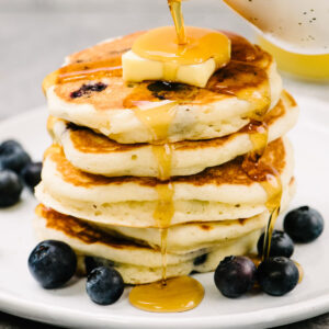 Side view, pouring maple syrup over a stack of blueberry buttermilk pancakes on a white plate, topped with a pat of butter and surrounded by additional fresh blueberries.