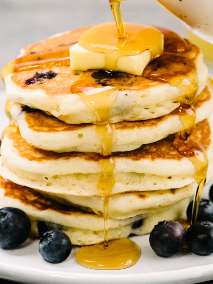 Side view, pouring maple syrup over a stack of blueberry buttermilk pancakes on a white plate, topped with a pat of butter and surrounded by additional fresh blueberries.