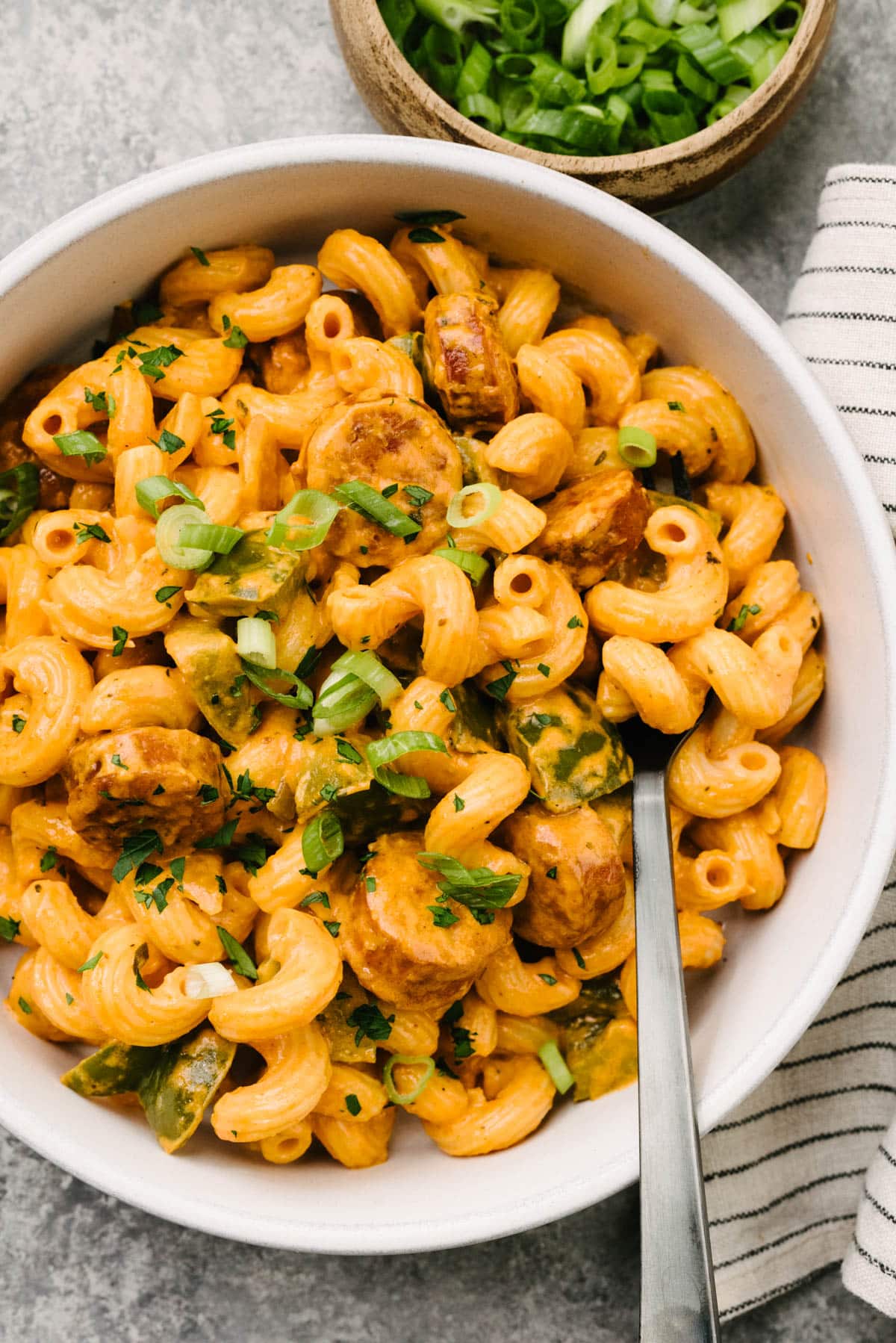 A fork tucked into a bowl of creamy cajun sausage pasta; a striped linen napkin and small bowl of sliced green onions surround the bowl.