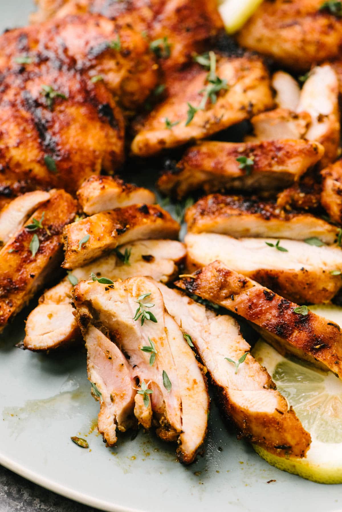 Side view, thinly sliced pieces of grilled chicken thigh on a light blue plate, garnished with fresh thyme and lemon slices.