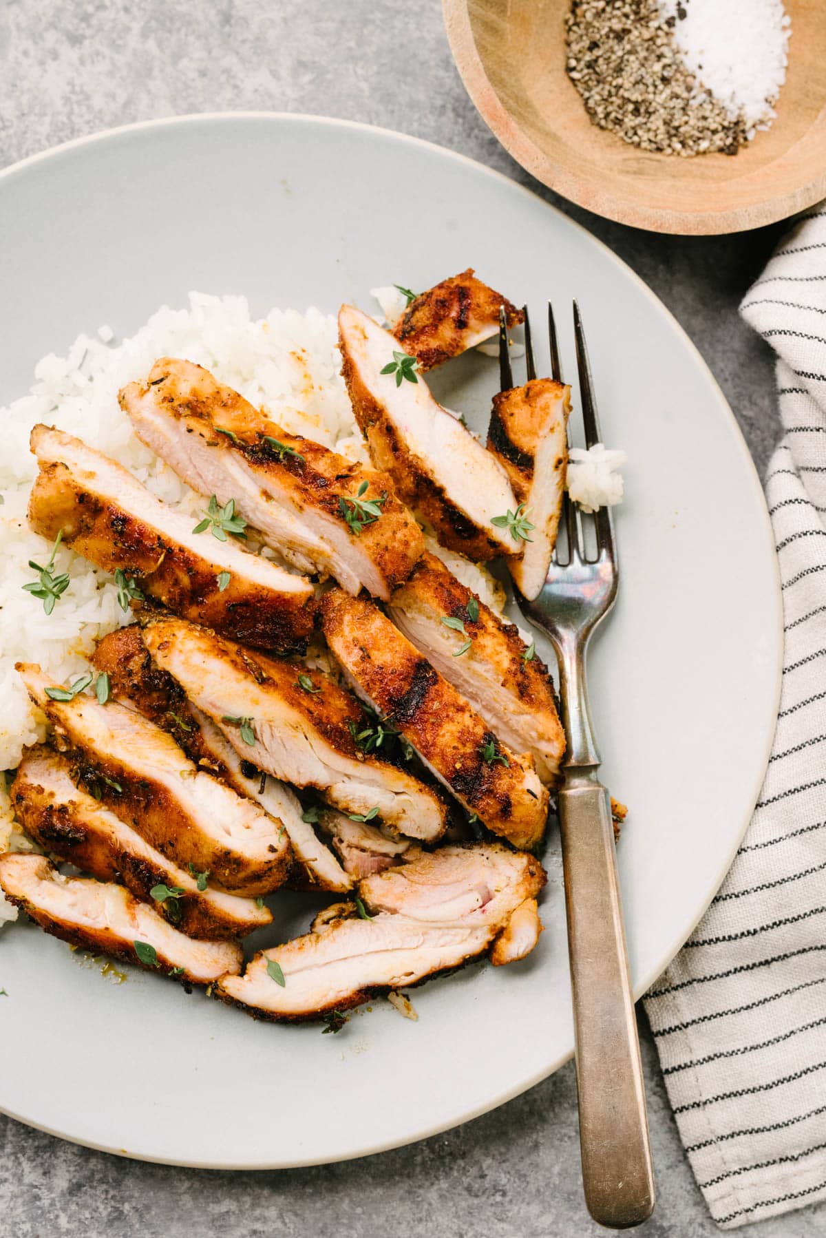 A fork tucked under sliced grilled chicken thighs over steamed white rice on a blue plate; a striped linen napkin and small bowl with salt and pepper are to the side.