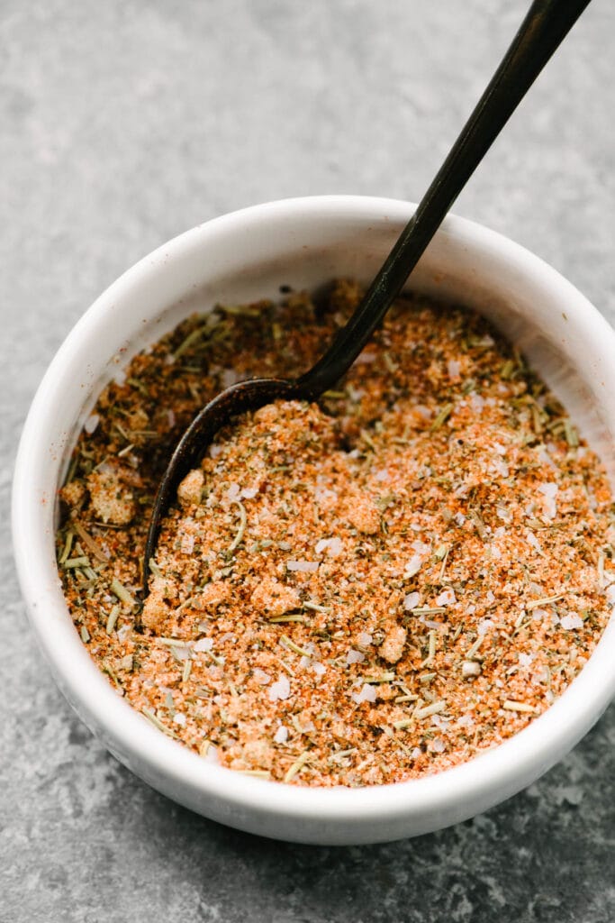 Side view, a spoon tucked into a small bowl of dry rub seasoning for boneless skinless chicken thighs.