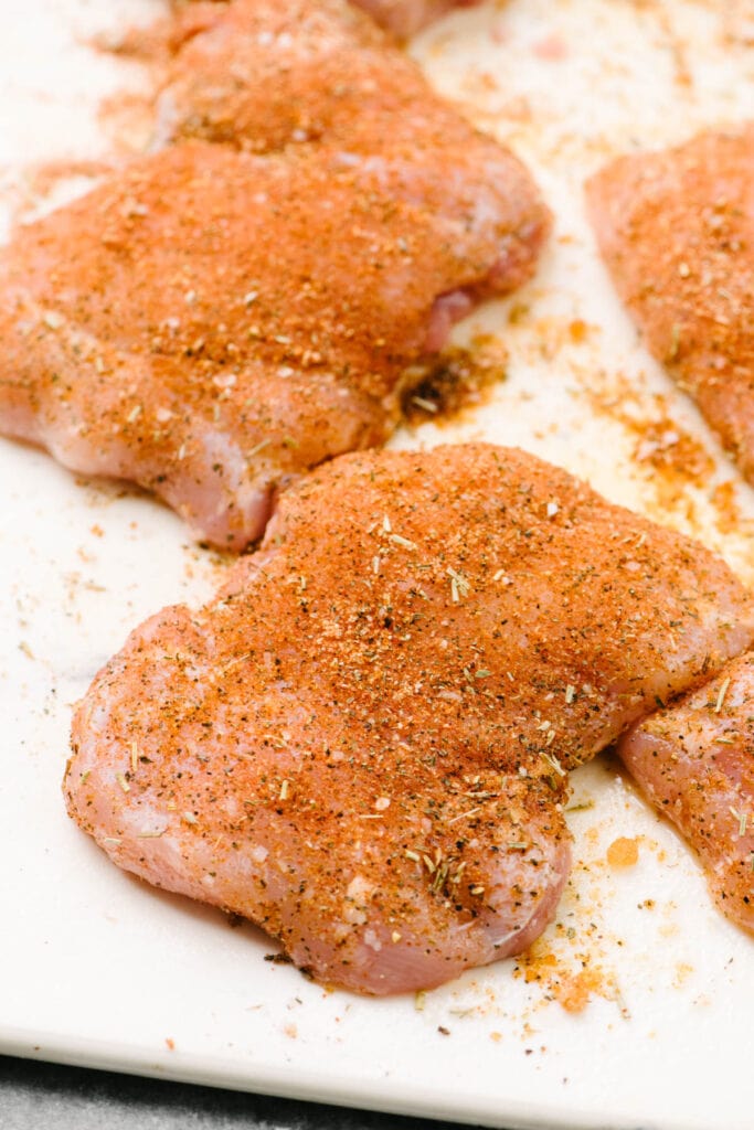 Side view, boneless skinless chicken thighs on a cutting board coated in olive oil and dry rub.