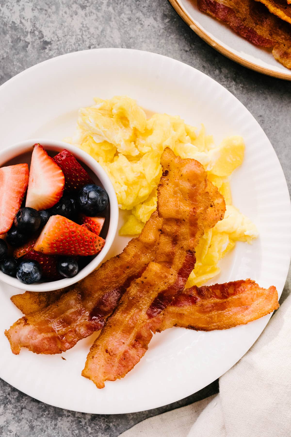 Crispy bacon, scrambled eggs, and a bowl of mixed berries on a white plate with a linen napkin to the side.