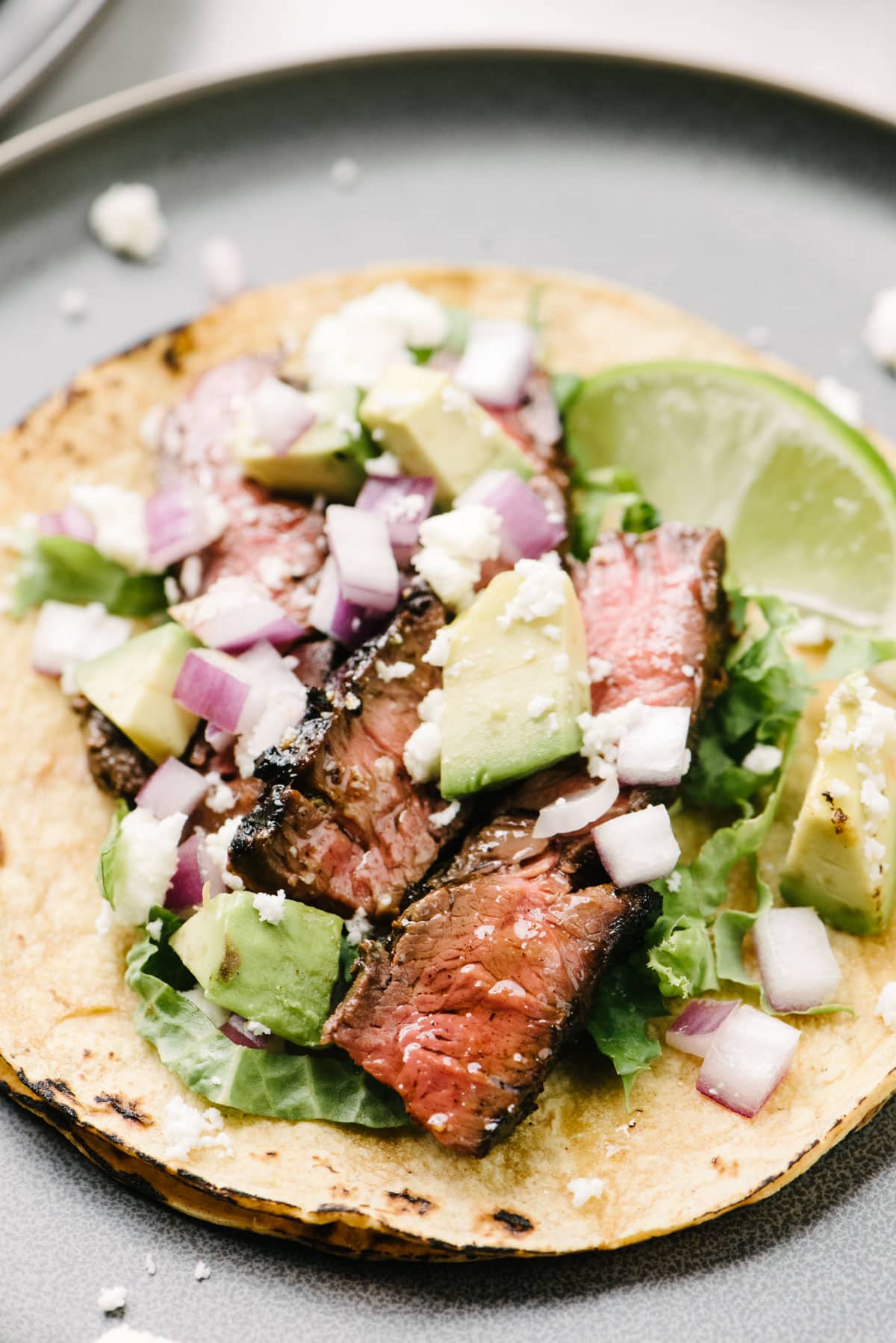 Side view, thin slices of skirt steak tucked into a corn tortilla and topped with avocado, red onion, and crumbled queso fresco cheese.