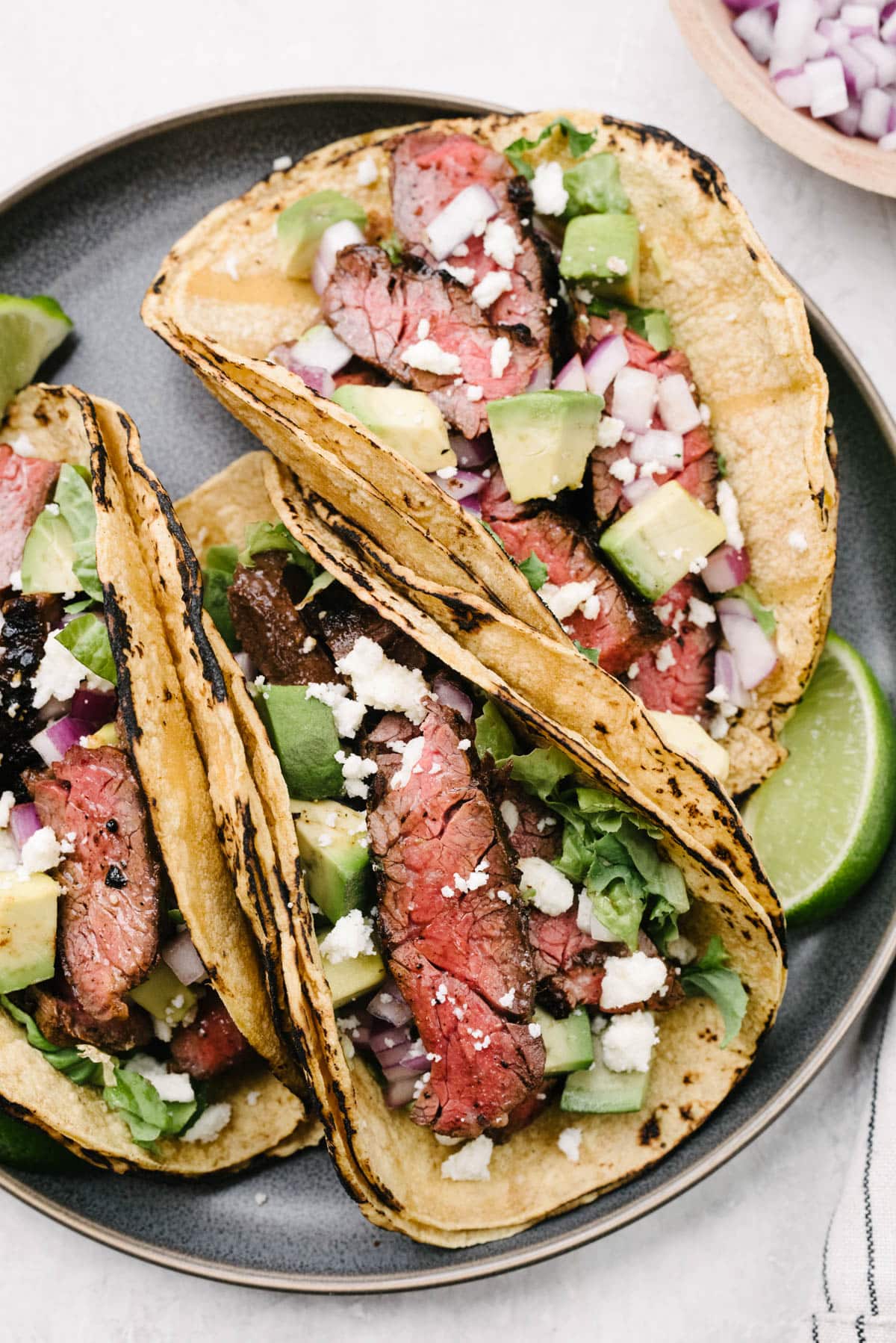 Three skirt steak tacos in corn tortilla shells on a grey plate, each topped with diced avocado, red onion, and crumbled queso fresco cheese.