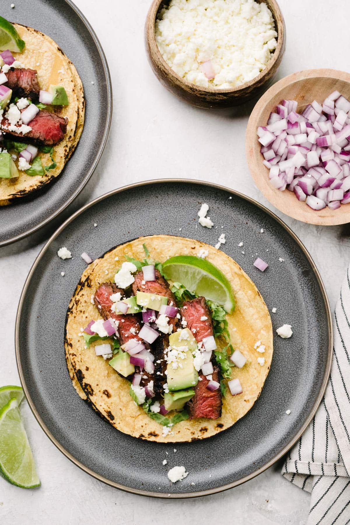 A skirt steak taco in a corn tortilla on a dark grey plate, topped with avocado, red onion, and queso fresco; the plate is surrounded by a striped linen napkin and small bowls of taco toppings.