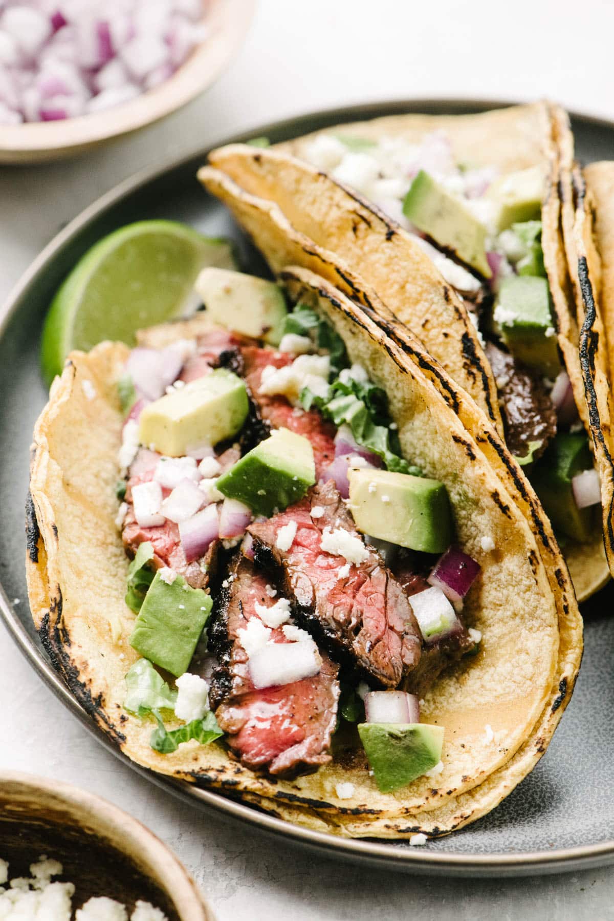 Side view, a close-up view of a steak taco in a corn tortilla shell with grilled skirt steak, avocado, red onion, and crumbled queso fresco cheese.