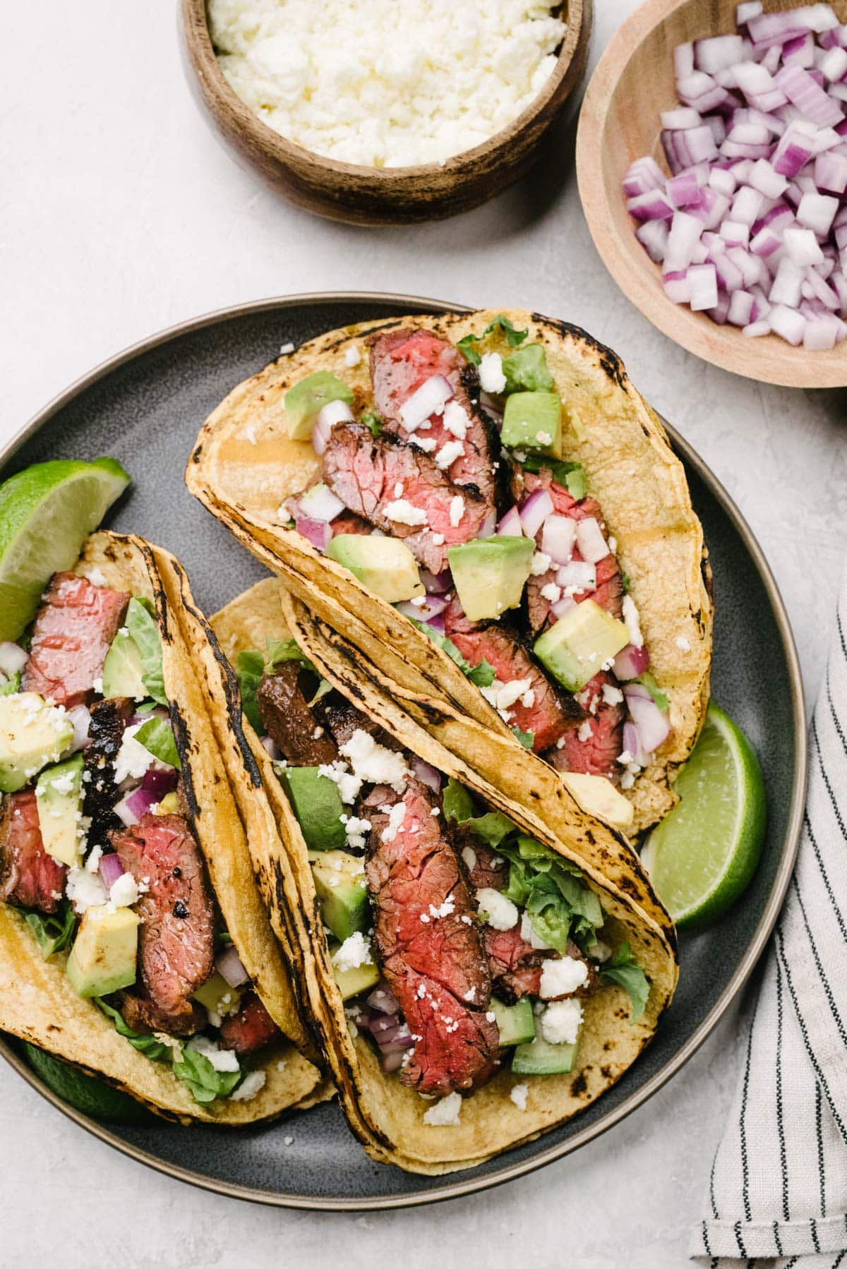 Three skirt steak tacos on a dark grey plate surrounded by a striped linen napkin and small bowls of queso fresco cheese and minced red onion.