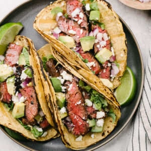 Three skirt steak tacos in corn tortilla shells on a grey plate, each topped with diced avocado, red onion, and crumbled queso fresco cheese.