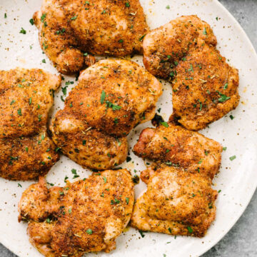 Baked boneless skinless chicken thighs on a speckled tan plate, garnished with fresh chopped parsley.