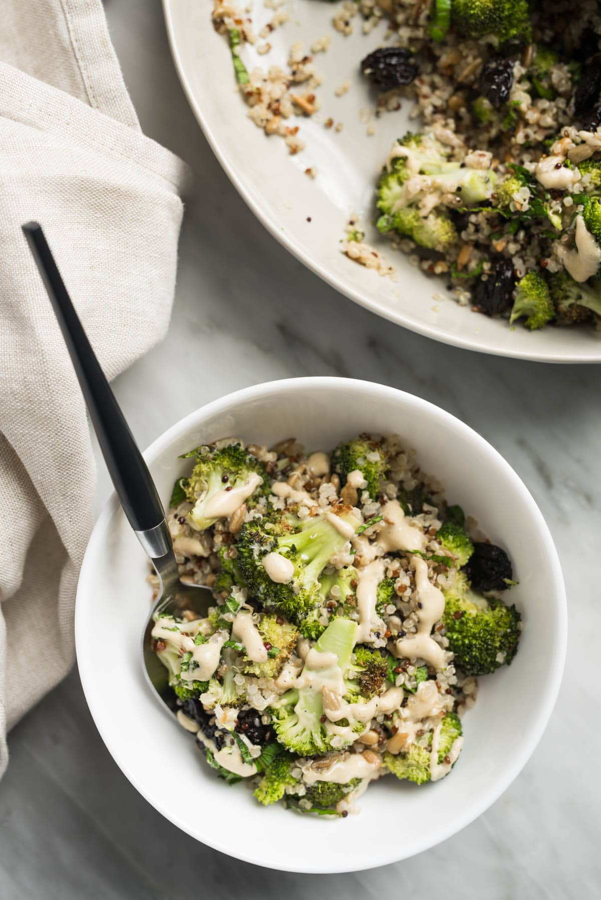 A spoon tucked into a small white bowl of broccoli quinoa salad on a marble table; a larger serving bowl and cream linen napkin surround the bowl.