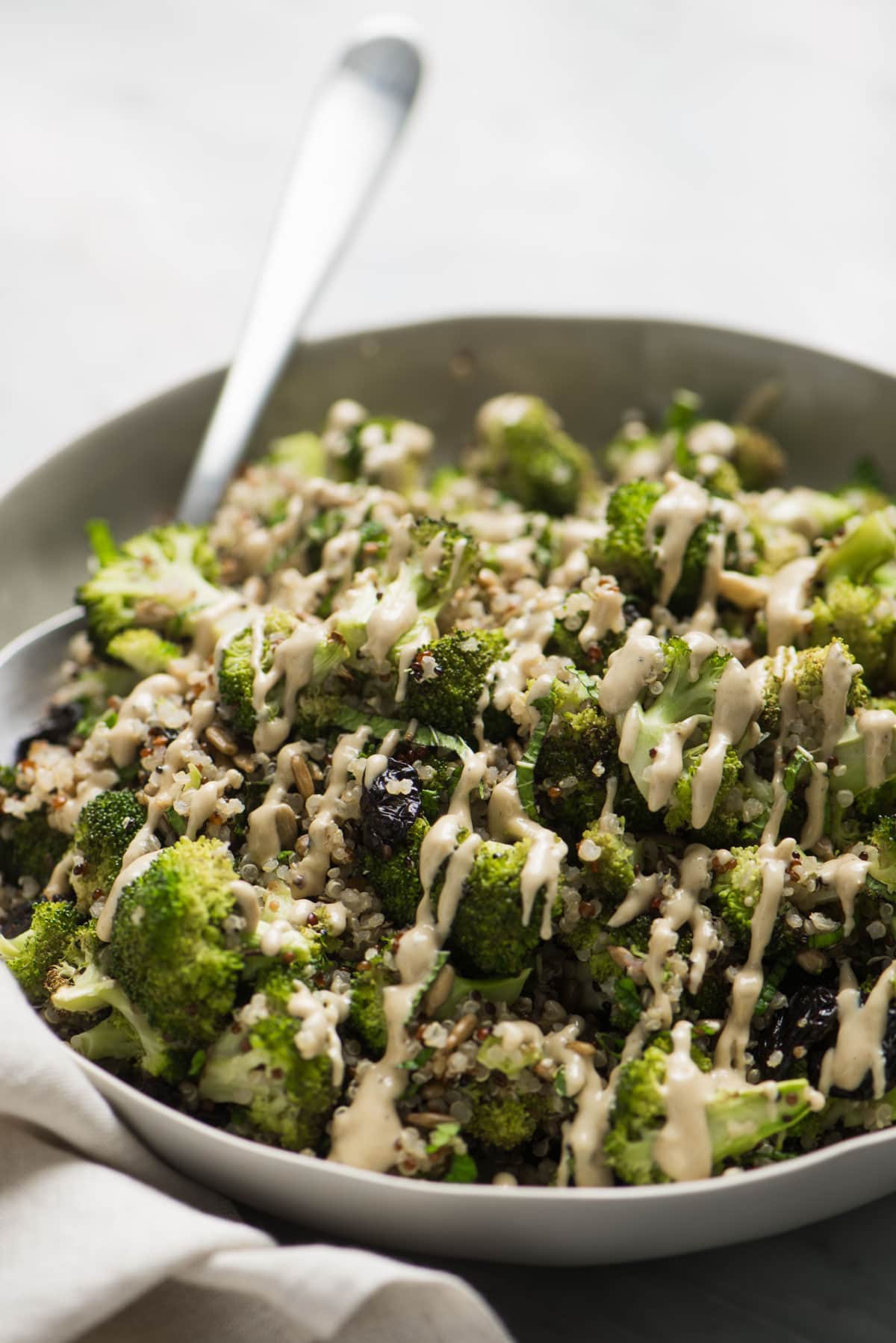 Side view, a serving spoon tucked into a low tan bowl filled with roasted broccoli and quinoa salad, drizzled with creamy sunflower seed butter dressing.