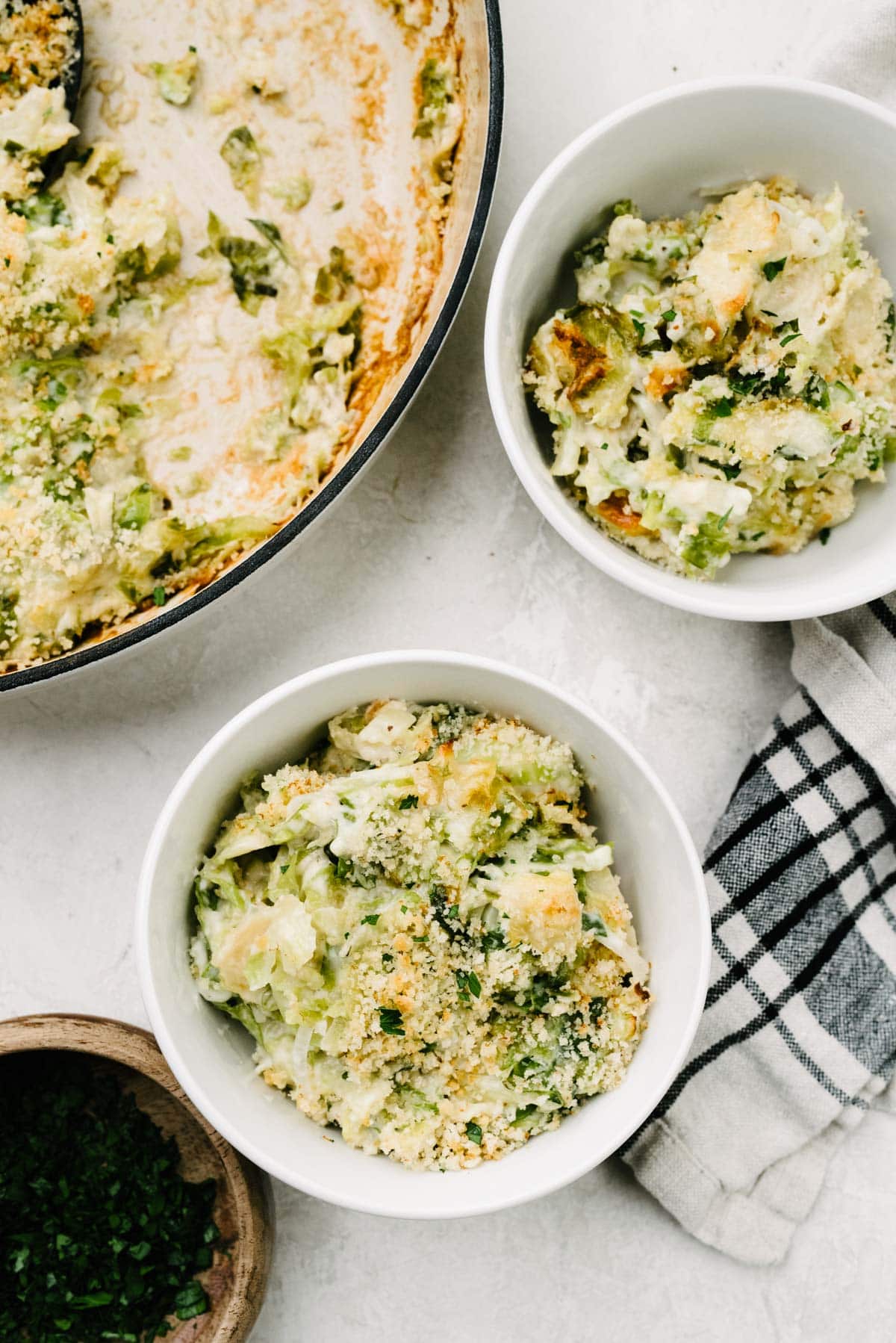 Two small white bowls filled with Brussels sprouts au gratin; a casserole dish, linen napkin, and small wood bowl of parsley surround the bowls.