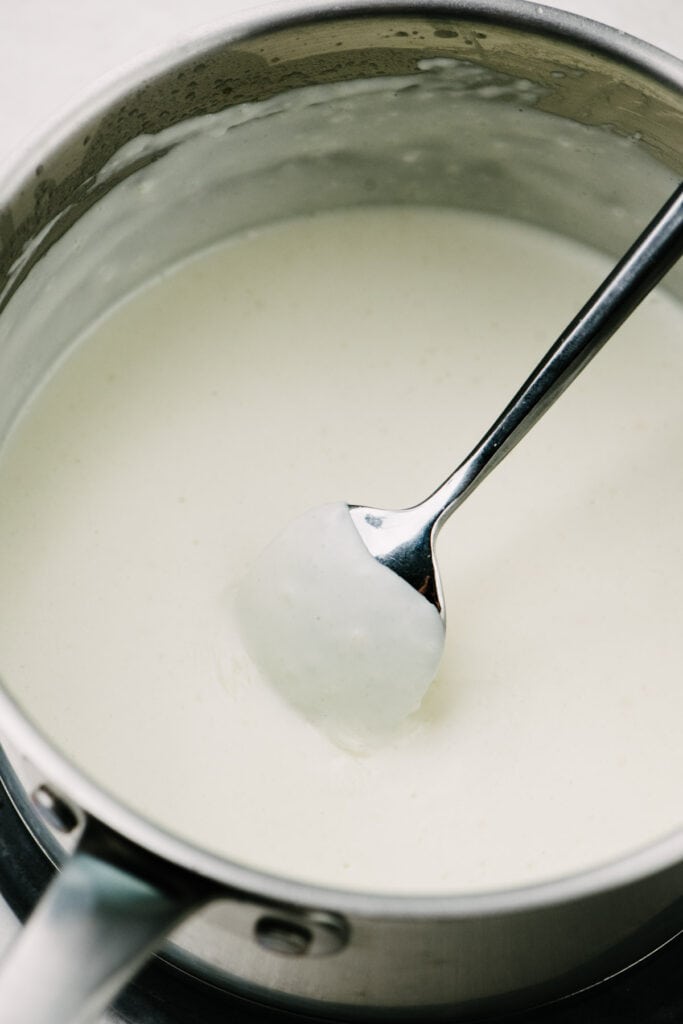 Side view, a spoon tucked into white sauce in a sauce pot showing that the sauce has thickened.