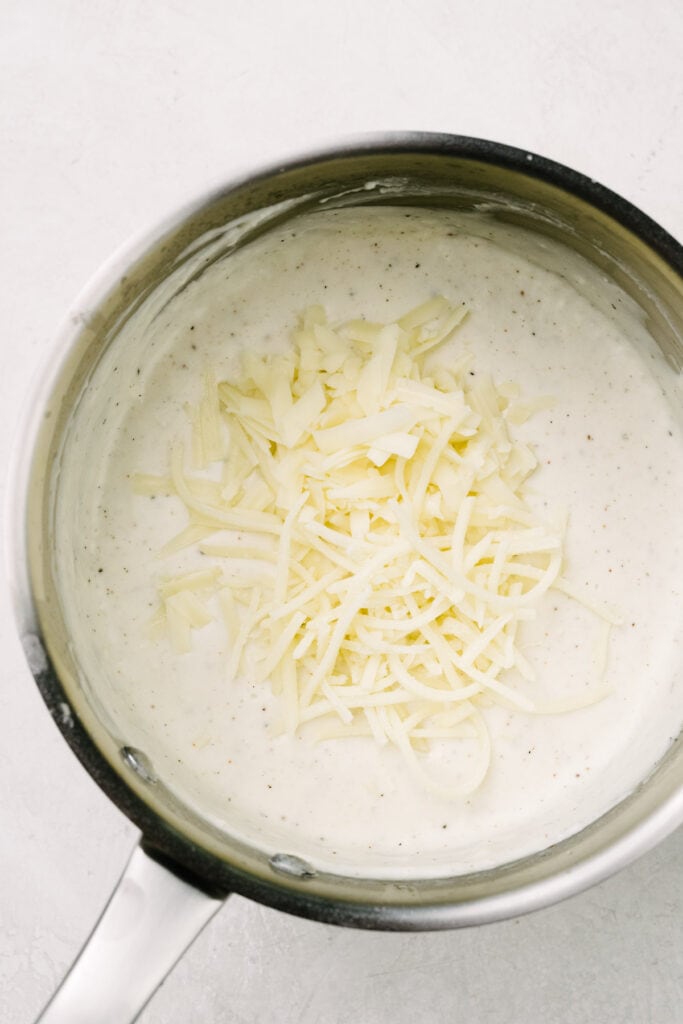 Adding shredded gruyere and sharp white cheddar cheeses to white sauce in a sauce pot.