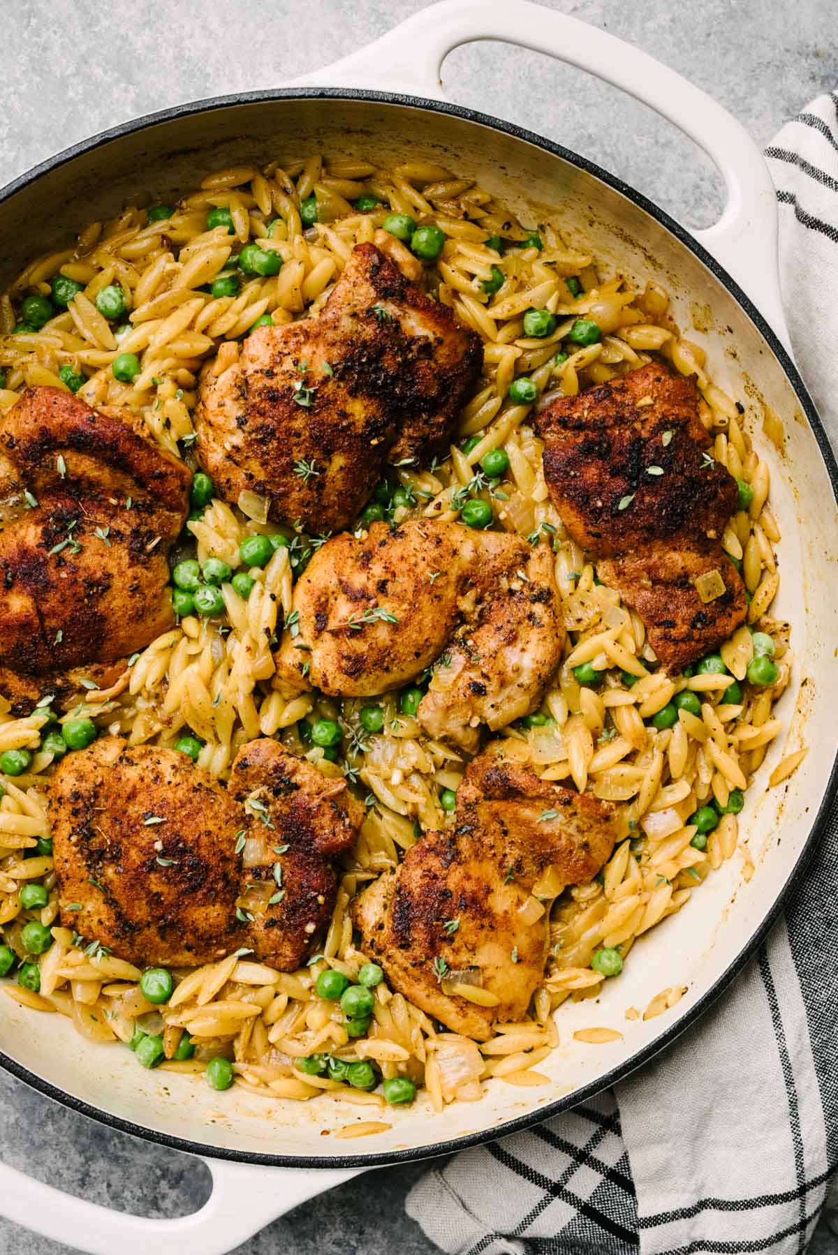 Seared chicken thighs over orzo pasta with peas in a large grey skillet with a striped linen napkin to the side.