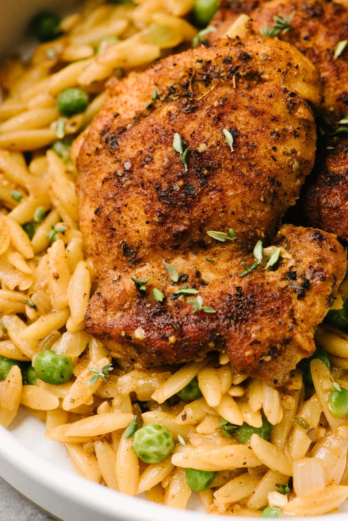 Side view, detail of a seared chicken thigh nestled into orzo pasta with peas in a low white dinner bowl.