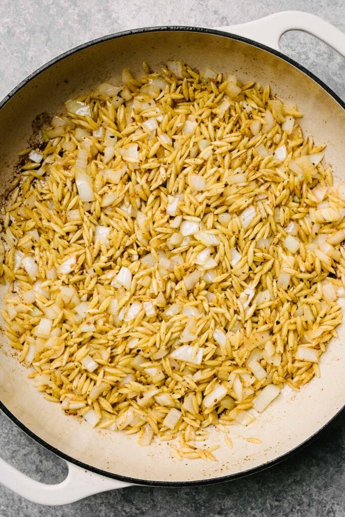 Toasted orzo pasta with onions and garlic in a large grey enameled skillet.