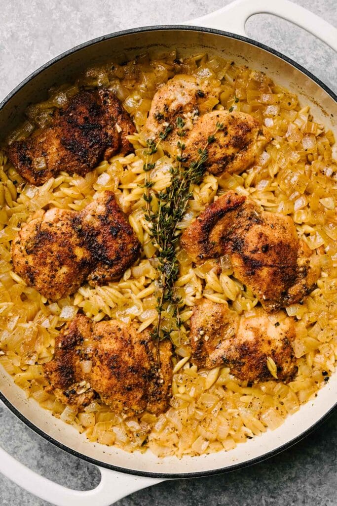 Cooked chicken thighs with orzo pasta and fresh thyme in a large grey skillet.