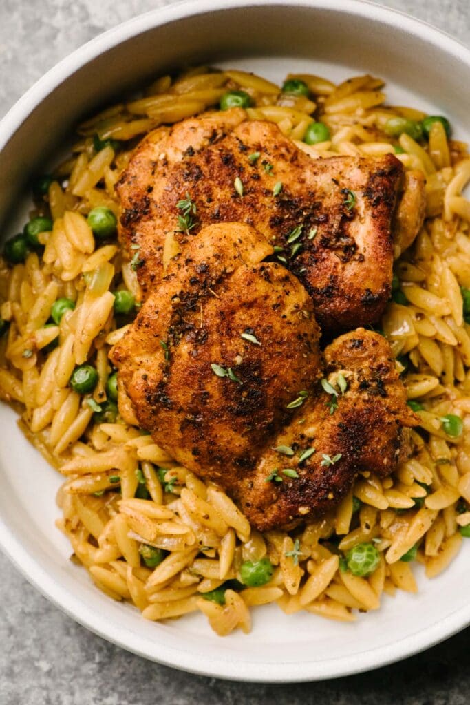 Seared chicken thighs over creamy orzo pasta with peas in a low white dinner bowl.