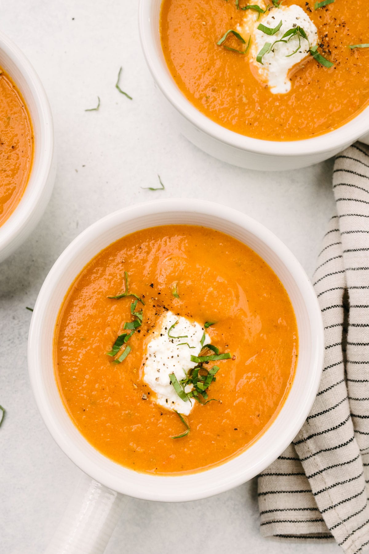 Three bowls of fresh tomato soup on a concrete background; each bowl is garnished with ricotta cheese and fresh basil, with a striped linen napkin tucked to the side.