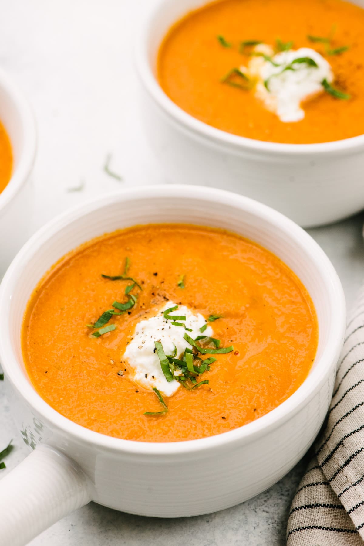 Side view, creamy tomato basil soup in several white bowls with a striped linen napkin to the side.