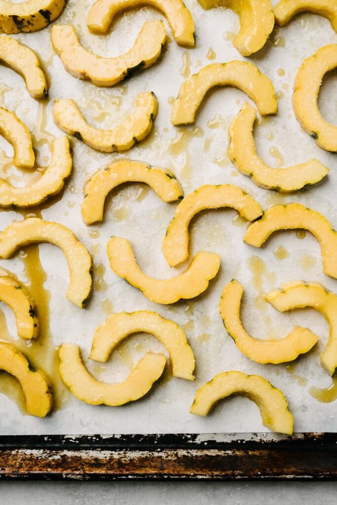 Delicata squash slices tossed with maple syrup and butter and sprinkled with salt on a parchment lined baking sheet.