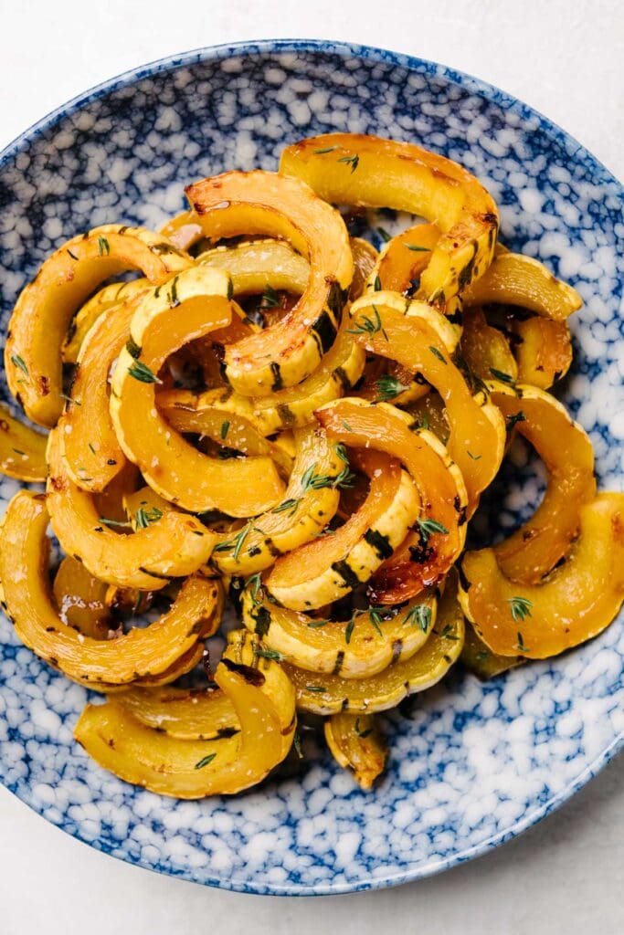 Roasted delicata squash slices with maple butter and fresh thyme in a blue speckled bowl.
