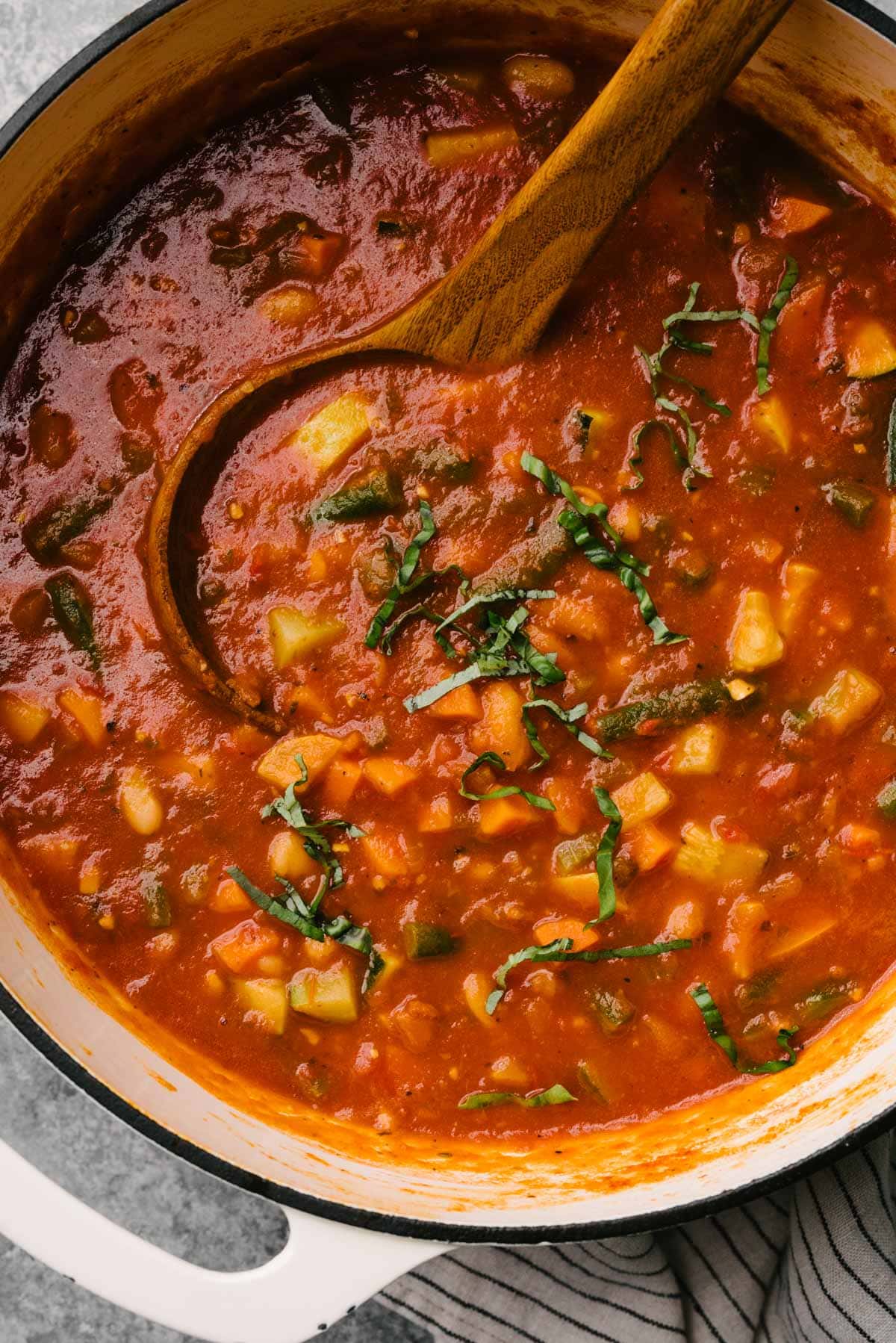 A wood ladle tucked into a soup pot of Italian vegetable soup, garnished with fresh basil.