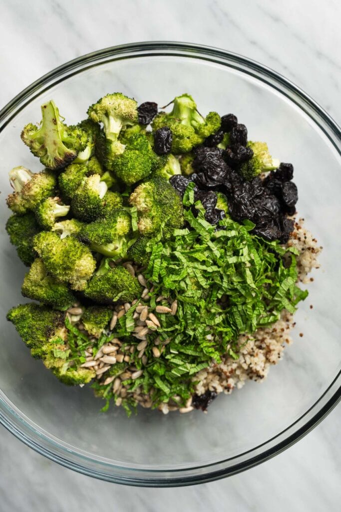 Cooked quinoa in a large glass mixing bowl, topped with roasted broccoli, dried cherries, sunflower seeds, and fresh mint.