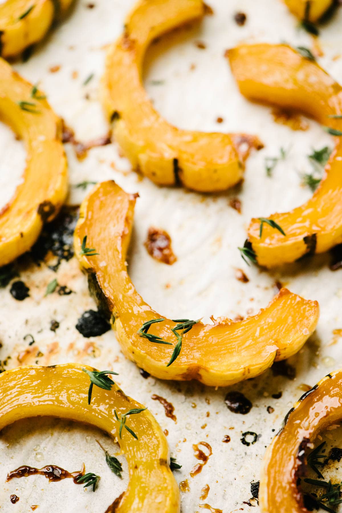 Side view, roasted delicata squash slices tossed with maple butter and fresh thyme on a parchment lined baking sheet.