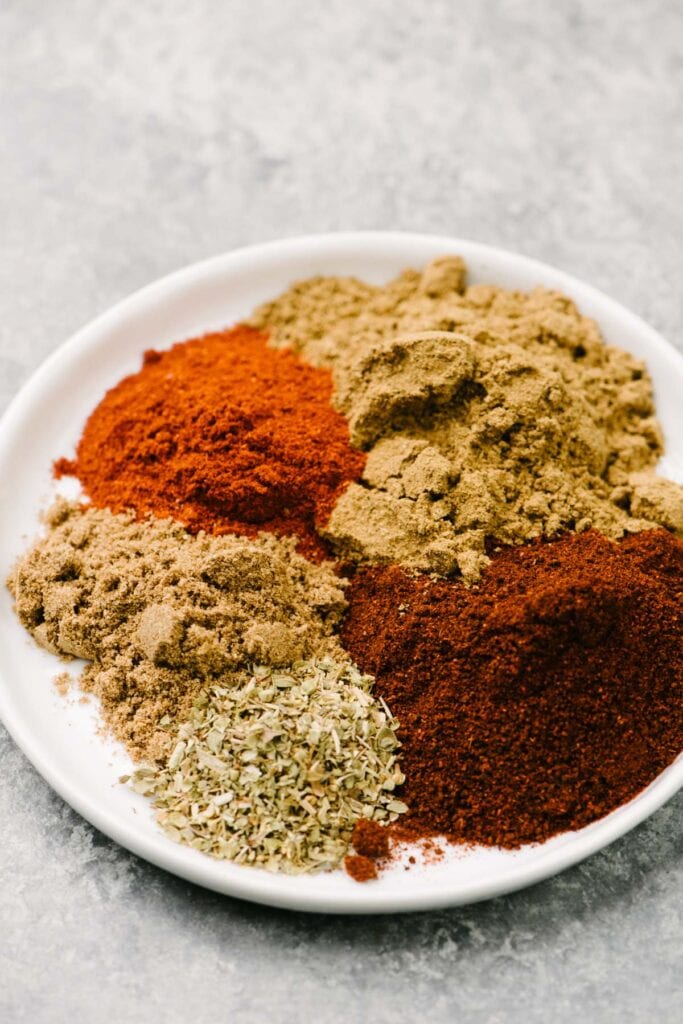 Side view - chili powder, cumin, paprika, coriander, and oregano on a small white plate.