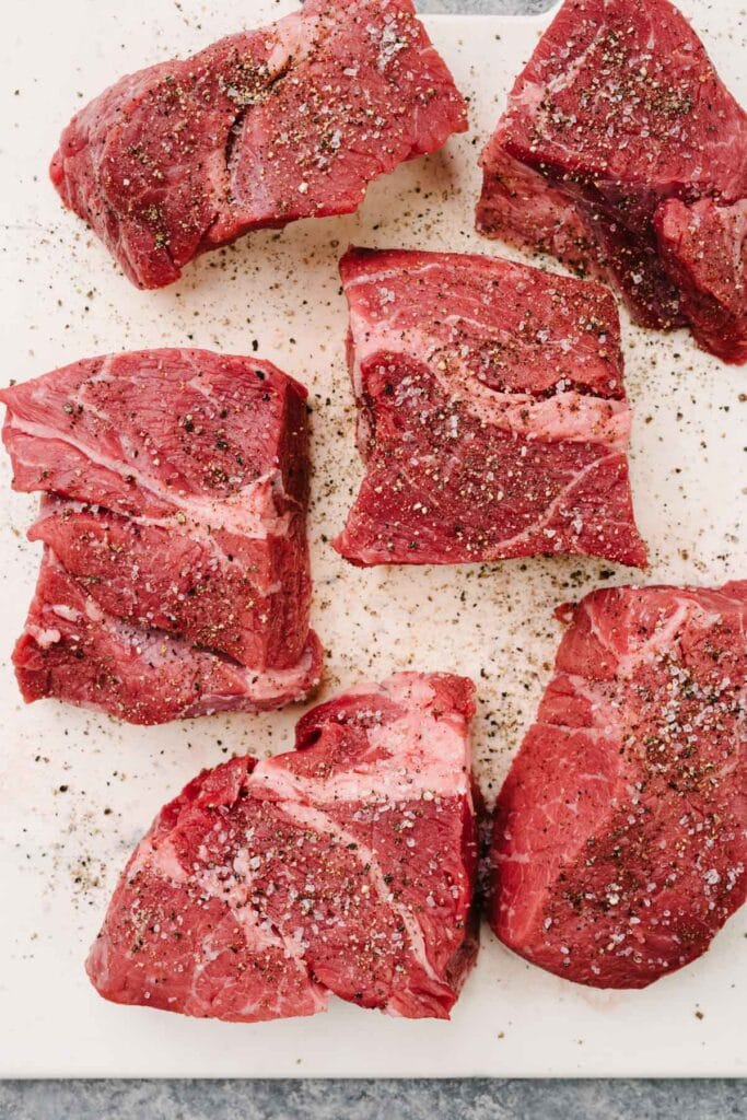 Whole chuck roast divided into six pieces and seasoned with salt and pepper on a white cutting board.