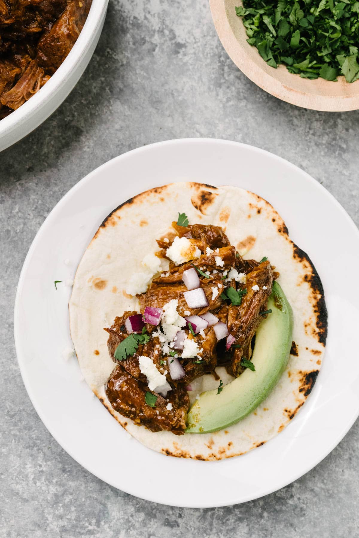 A flour tortilla on a white plate piled with Mexican shredded beef, avocado, red onion, queso freso, and cilantro. 