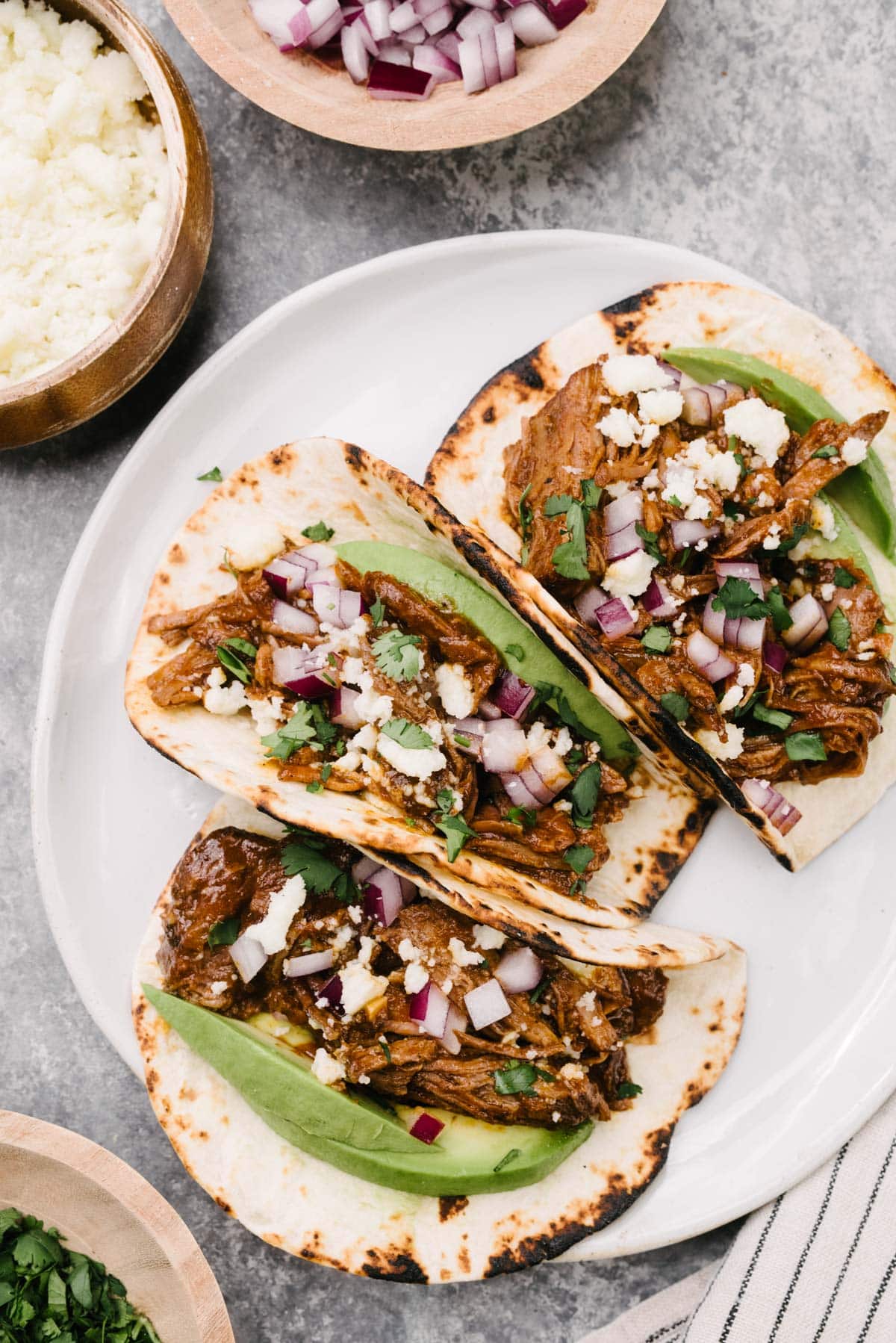 Three shredded beef tacos on a white plate garnished with avocado slices, diced red onion, and crumbled queso fresco; a linen napkin and small bowls of garnishes surround the plate.