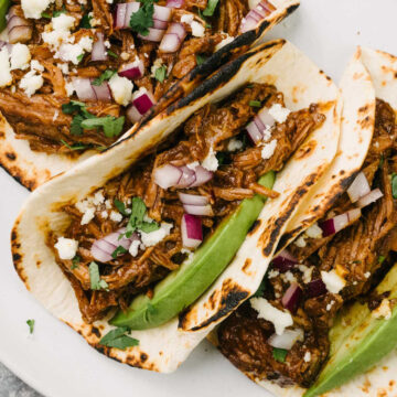 Three tacos on a plate filled with Mexican shredded beef and garnished with avocado, green onion, queso fresco, and fresh cilantro.