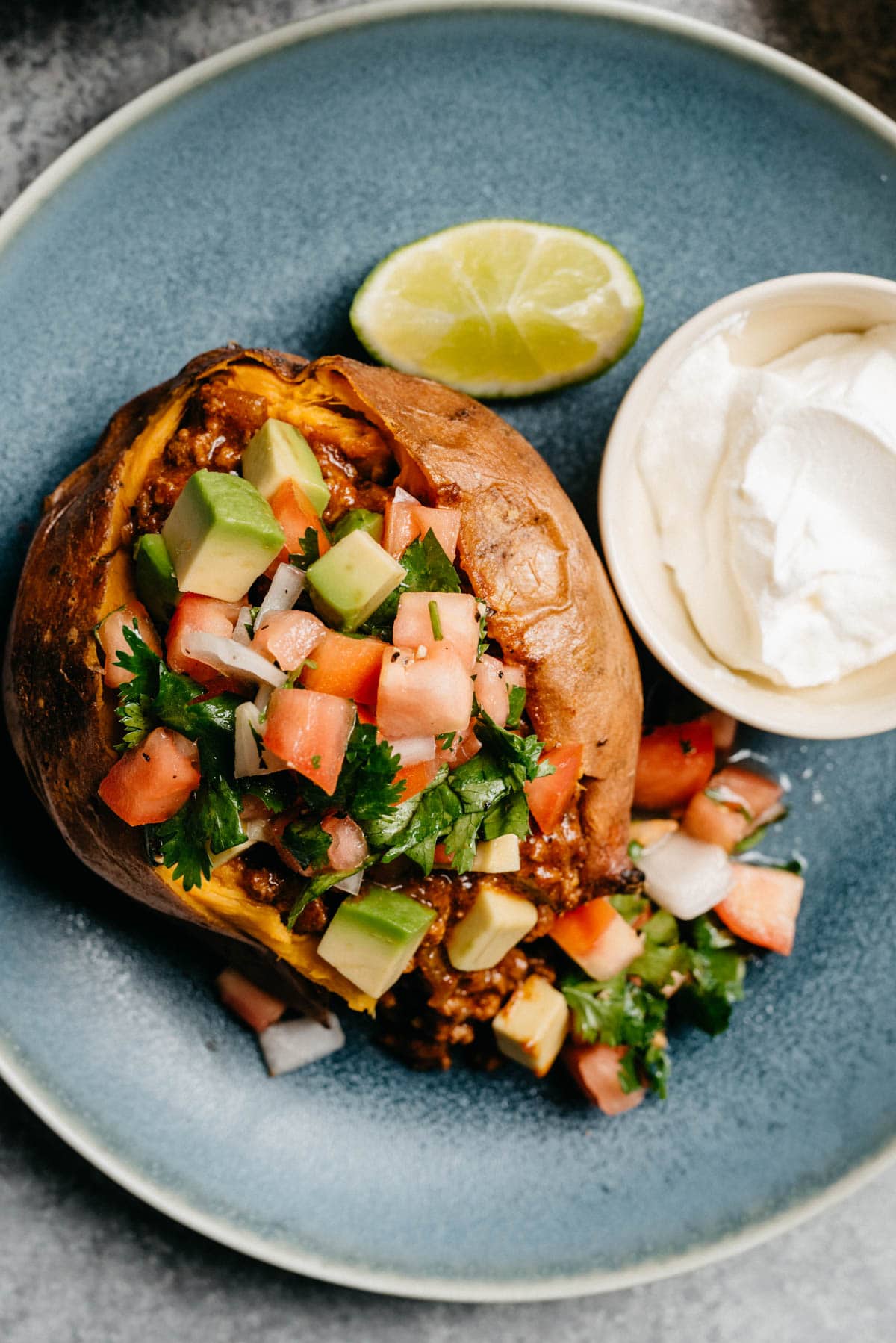 A taco stuffed sweet potato on a blue plate, topped with pico de gallo and avocado; a small dish of sour cream and a lime wedge are also on the plate.