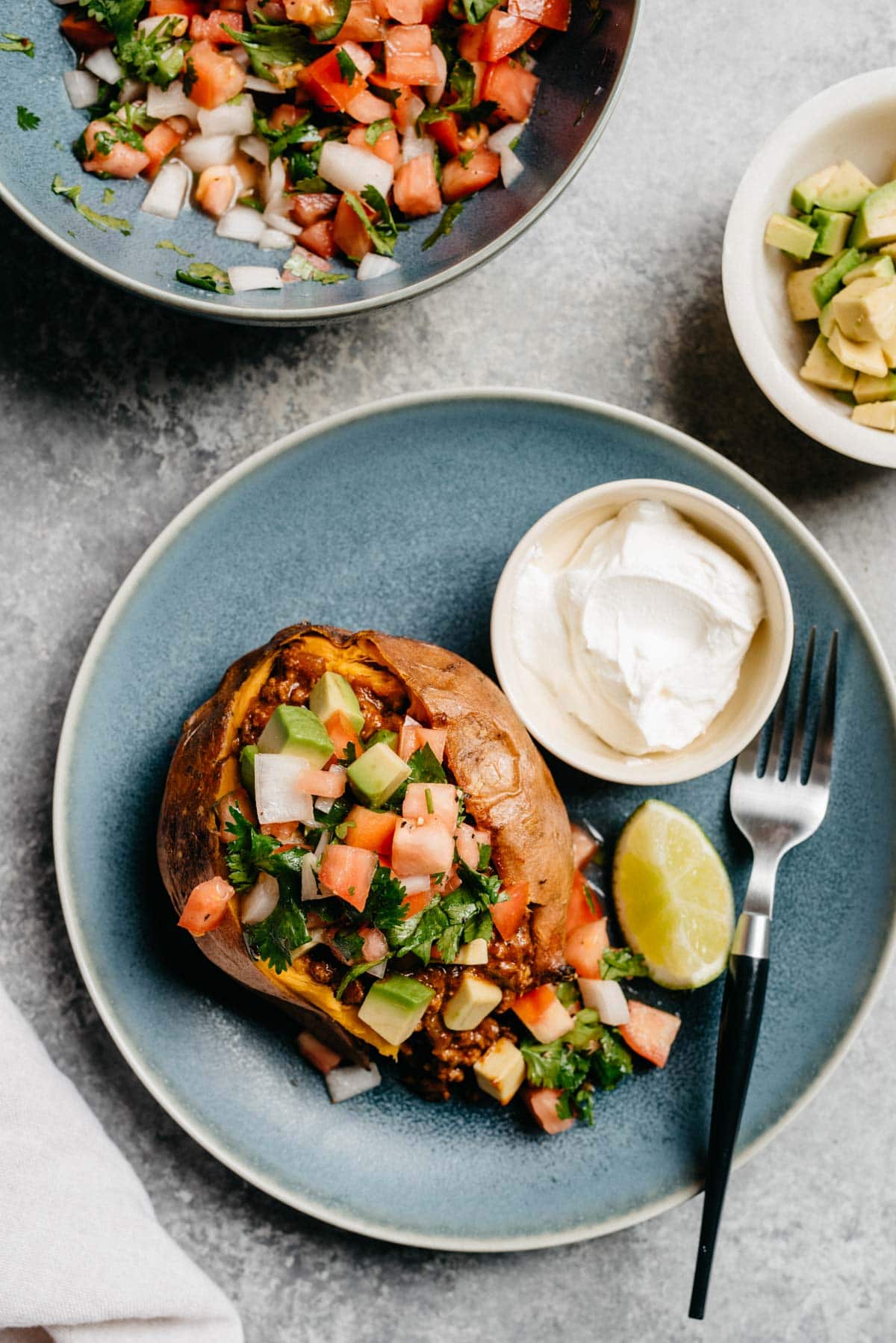 A baked potato stuffed with ground beef taco meat on a blue plate; small bowls of pico de gallo and avocado to the side, along with a linen napkin.
