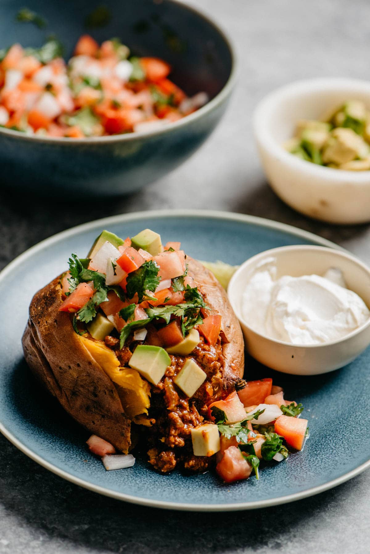 Side view, a taco stuffed sweet potato on a blue plate, garnished with pico de gallo and avocado; small bowls of various toppings are in the background.