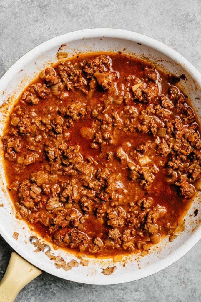 Tomato paste and ground beef poured into a skillet with sauteed ground beef, onions, and taco seasoning.