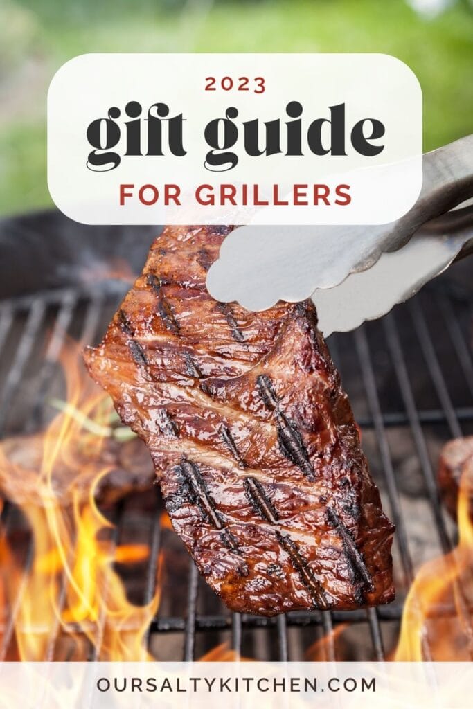Side view, a steak being lifted from a grill by tongs, surrounded by flames; text overlay reads "2023 gift guide for grillers".