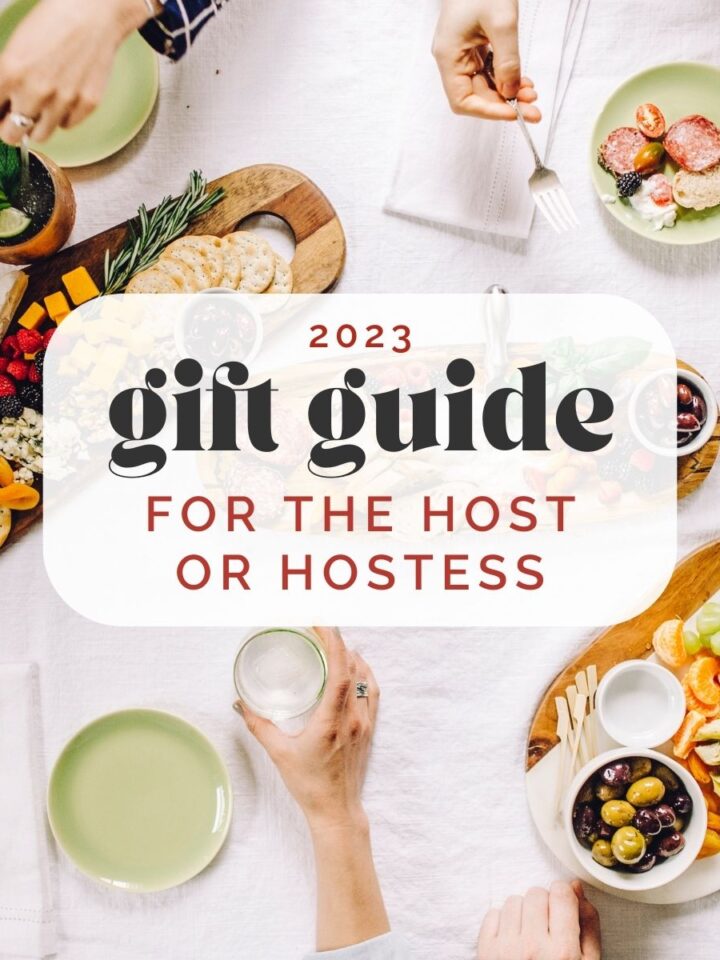 An overhead shot of a table arranged with various cheese and charcuterie platters, cocktails, and small plates arranged on a white linen tablecloth; a text overlay reads "2023 gift guide for the host or hostess".