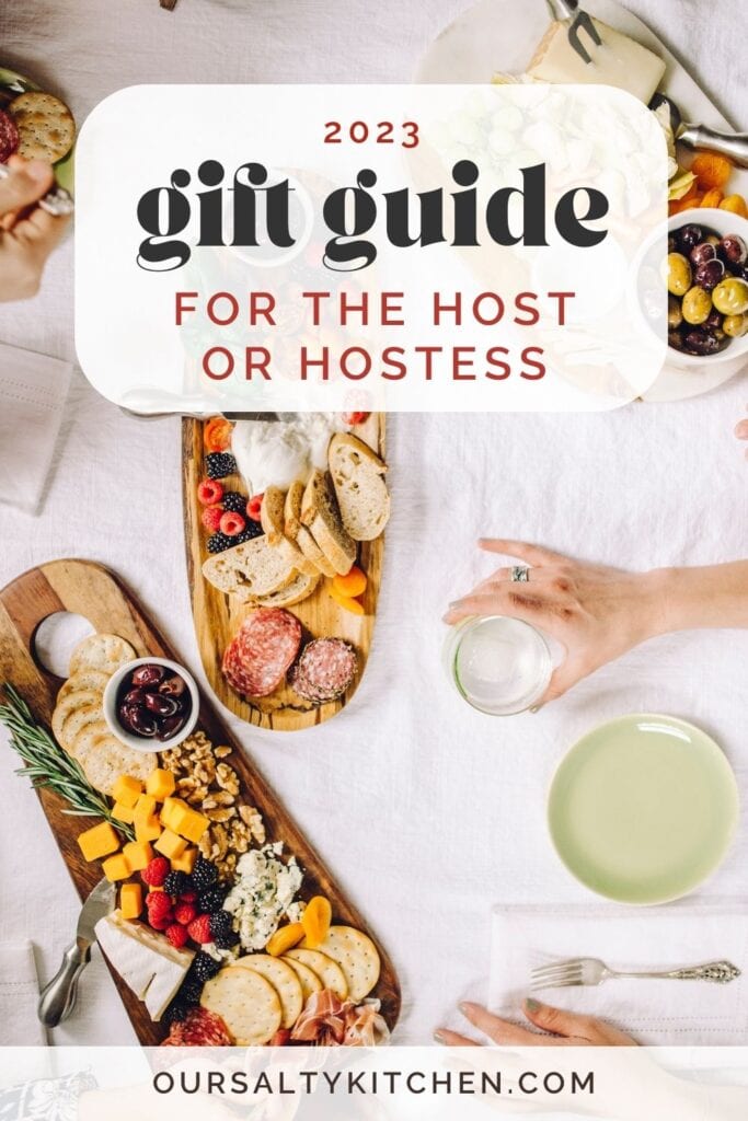 An overhead shot of a table arranged with various cheese and charcuterie platters, cocktails, and small plates arranged on a white linen tablecloth; a text overlay reads "2023 gift guide for the host or hostess".